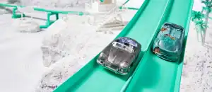 Miniature toy cars racing down a green slide in a playful outdoor setting.