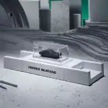 Miniature black Mustang car model in a clear display case on a white platform.