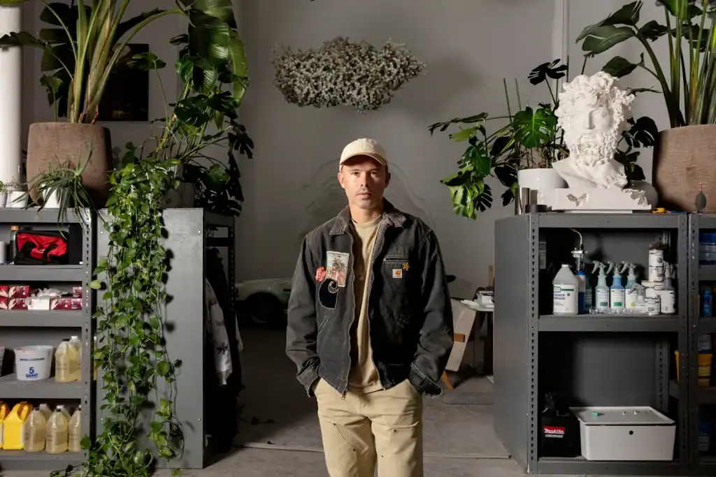 Daniel Arsham Man standing in a warehouse surrounded by shelves with art supplies and plants.